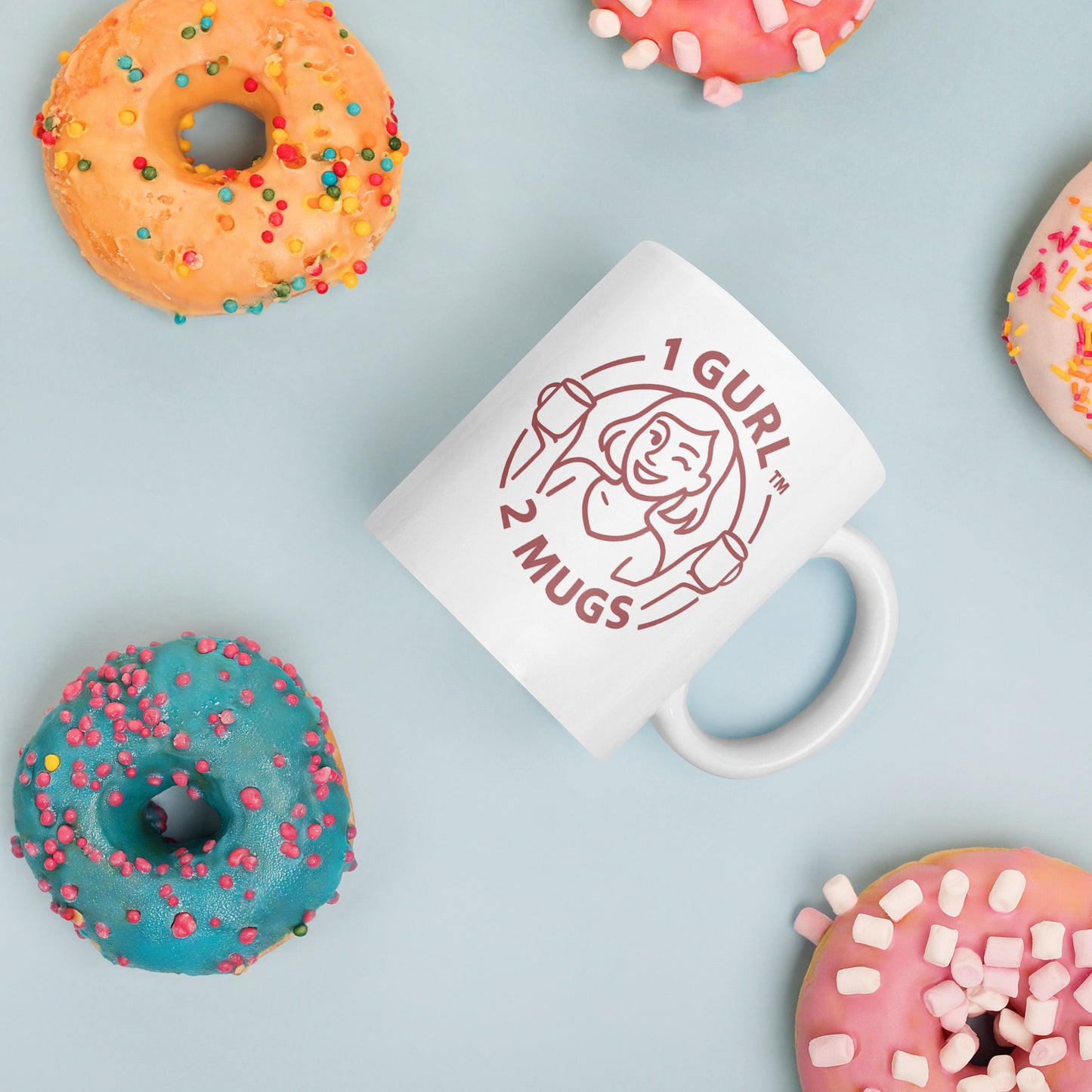 White mug with '1 Gurl 2 Mugs' design surrounded by colorful donuts on a light blue background