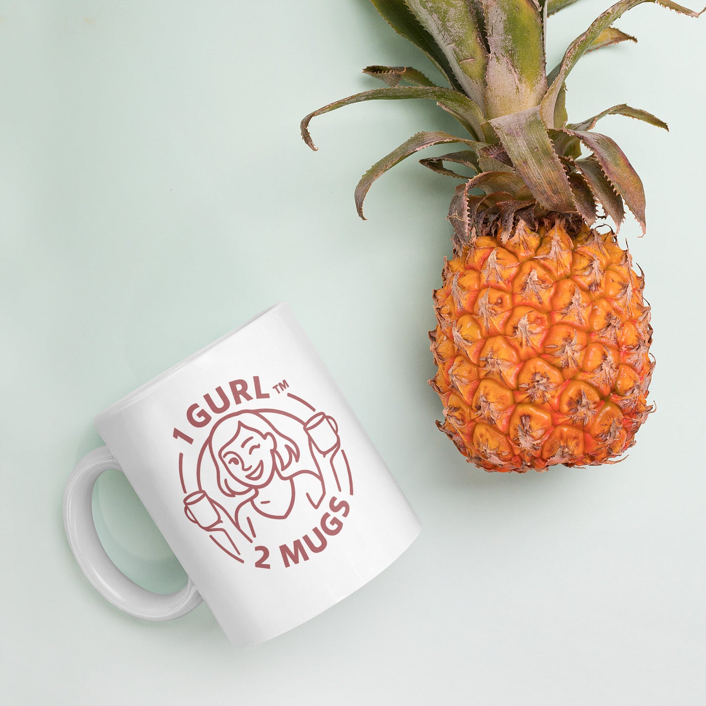 White mug with '1 Gurl 2 Mugs' design next to a pineapple on a light green background