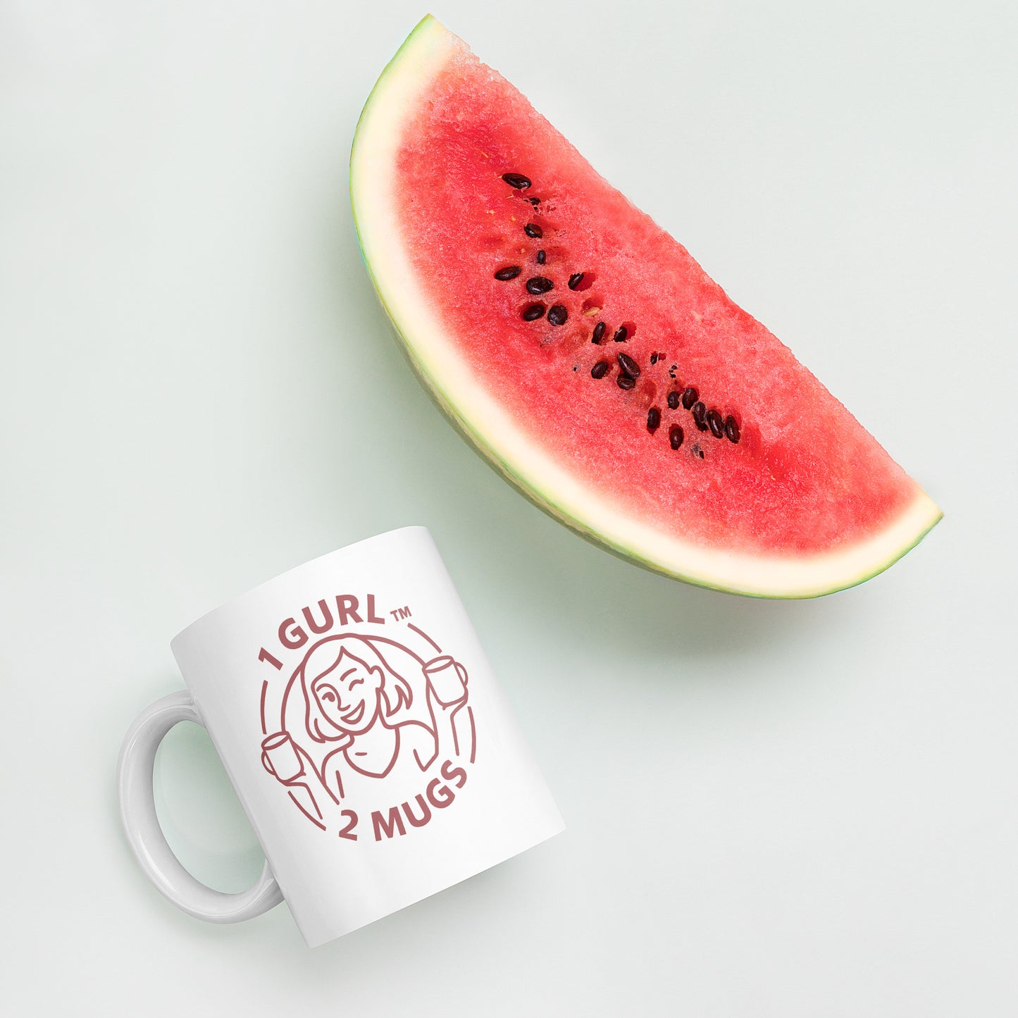 White mug with '1 GURL 2 MUGS' design next to a slice of watermelon on a light background