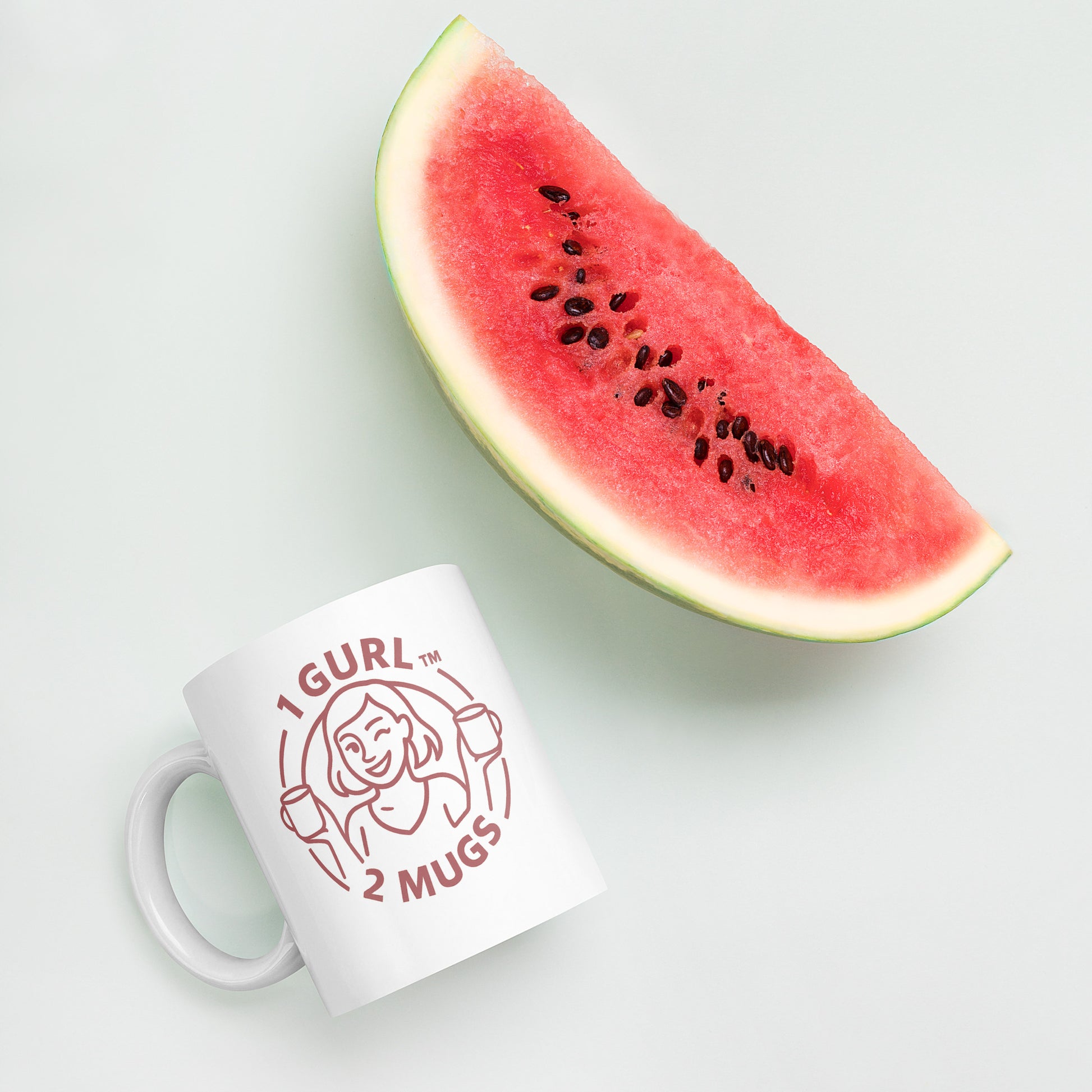 White mug with '1 GURL 2 MUGS' design next to a slice of watermelon on a light background